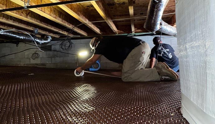 Two workers installing a dimpled drainage mat in a crawlspace