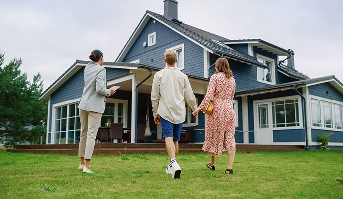 A professional realtor shows a young couple the exterior of a large, blue, two-story house on a grassy lawn.